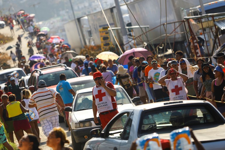 An Acapulco street is crowded with cars and a long line of people, stretching toward the horizon, as aid workers — visible with a white apron emblazoned with a red cross — stand watching.